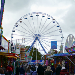 Ein beleuchtetes Riesenrad mit Gondeln dreht sich im Hintergrund, während im Vordergrund Menschen spazieren. Geschäfte mit ansprechender Beleuchtungstechnik säumen den Weg und schaffen eine einladende Atmosphäre